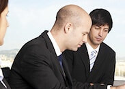 Drei Geschäftsleute diskutieren, wobei ein Mann im Mittelpunkt steht und nachdenklich nach unten blickt; seine Stimme ist kaum zu hören.