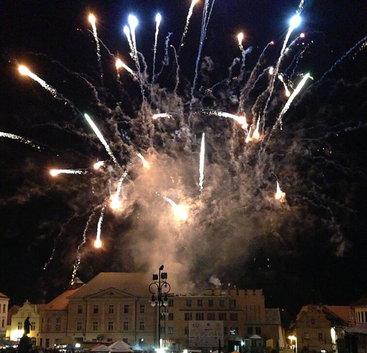 Über einem Stadtplatz explodieren nachts Feuerwerke und ihr Klang erleuchtet die Fassade eines historischen Gebäudes.