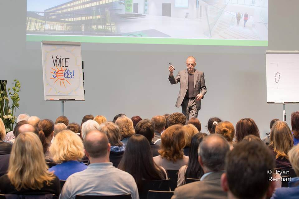 Ein Redner im Anzug hält auf einer Konferenz einen Vortrag vor einem Publikum und nutzt dabei visuelle Hilfsmittel, darunter Poster und ein projiziertes Bild.