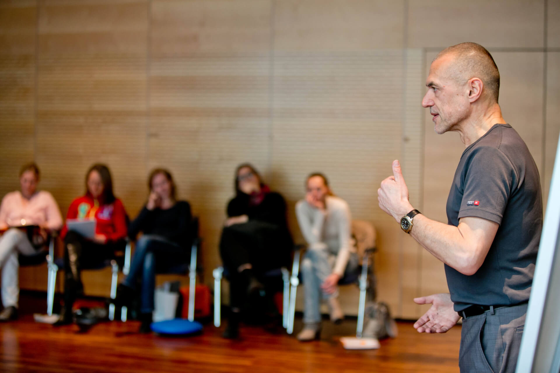 Ein Personal Trainer hält mit einer Daumen-hoch-Geste eine Präsentation vor einem aufmerksamen Publikum, das in einem Raum mit Holzhintergrund auf dem Boden sitzt.