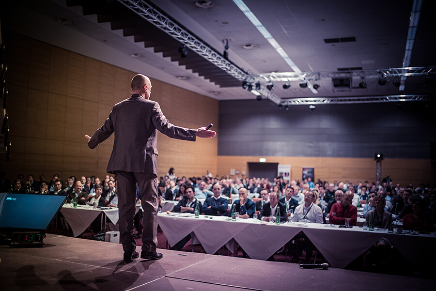 Ein Mann hält auf der Bühne über einen Lautsprecher einen Vortrag vor einem Publikum in einem Konferenzsaal.