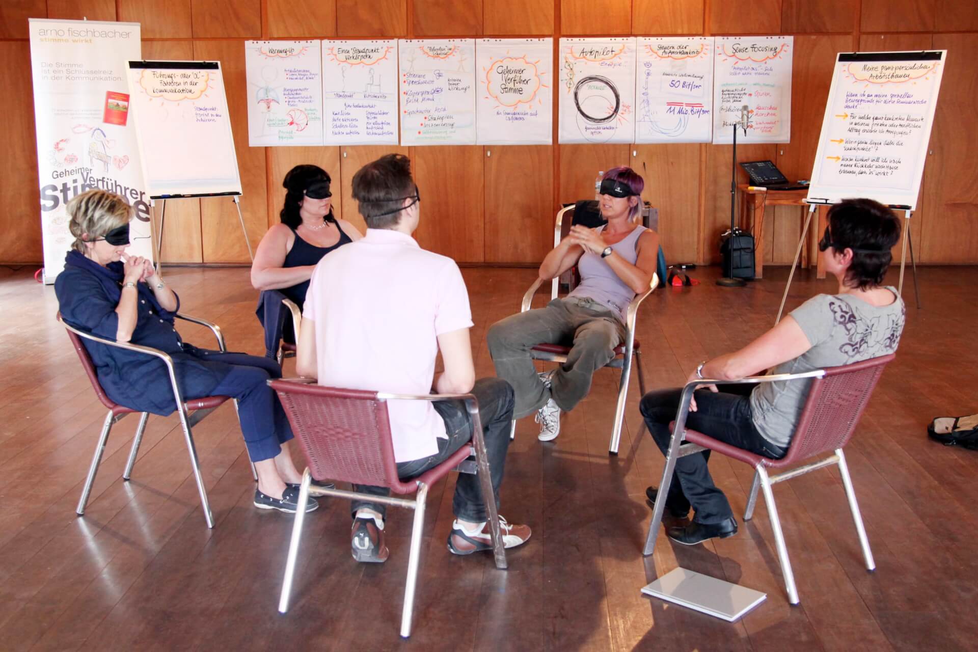 Fünf Erwachsene sitzen auf Stühlen im Kreis, einer fungiert als Sprecher. Sie diskutieren in einem hell erleuchteten Raum, um sie herum sind Papiere und Whiteboards voller Notizen verteilt.