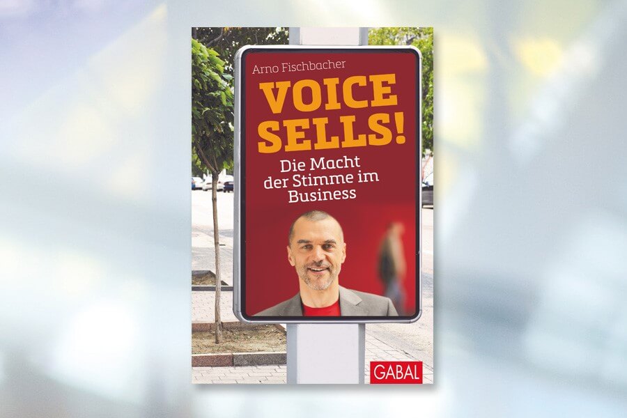 Werbe-Straßenbanner mit dem Buch „Macht der Stimme im Business“ von Arno Fischbacher, ausgestellt auf einem Bürgersteig in der Stadt mit unscharfem Hintergrund.