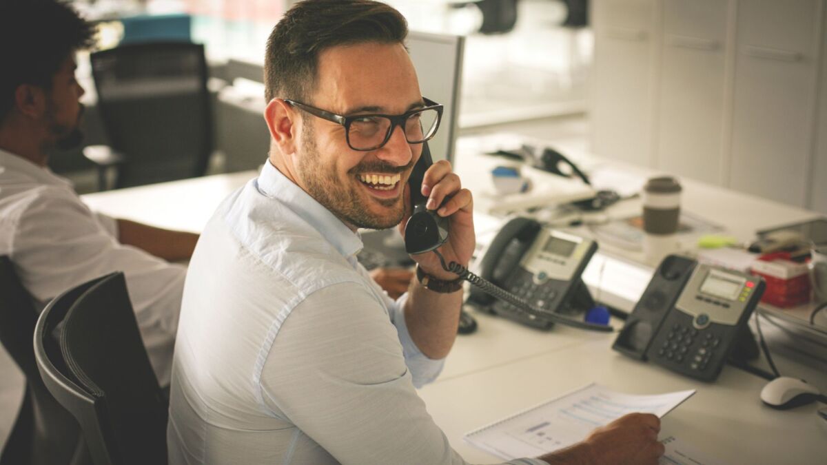 Ein Mann mit Brille lächelt, während er an seinem Schreibtisch in einer Büroumgebung Outbound-Telefonie betreibt. Im Hintergrund sitzt eine weitere Person an einem Schreibtisch. Auf dem Schreibtisch liegen mehrere Telefone und Papiere verstreut.