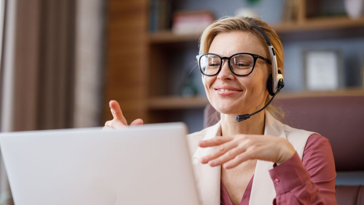 Eine Frau mit Brille und Headset lächelt und gestikuliert, während sie in einem gut beleuchteten Raum auf ihren Laptop schaut und offensichtlich in ein Outbound-Telefon vertieft ist.