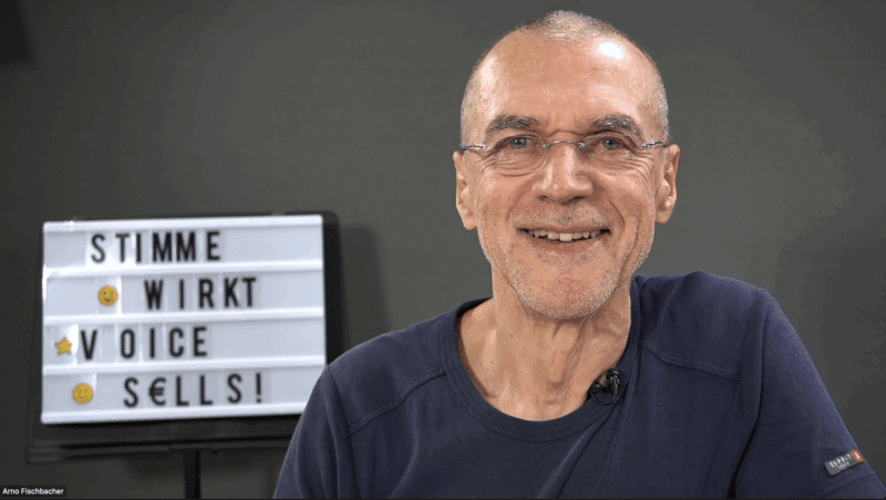 Ein Mann mit Brille lächelt in die Kamera. Hinter ihm steht auf einem Schild: „STIMME WIRKT VOICE SELLS!“ mit Emoji-Akzenten – perfekt für ein Seminar zum Thema Rhetorik für Führungskräfte.