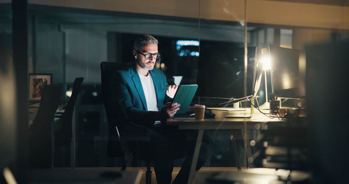 Ein Mann im Anzug arbeitet spätabends in einem modernen Büro, sitzt an einem Schreibtisch mit einem Tablet, das von einer Schreibtischlampe beleuchtet wird - vielleicht bereitet er seine Präsentation "Rhetorik für Führungskräfte" vor.