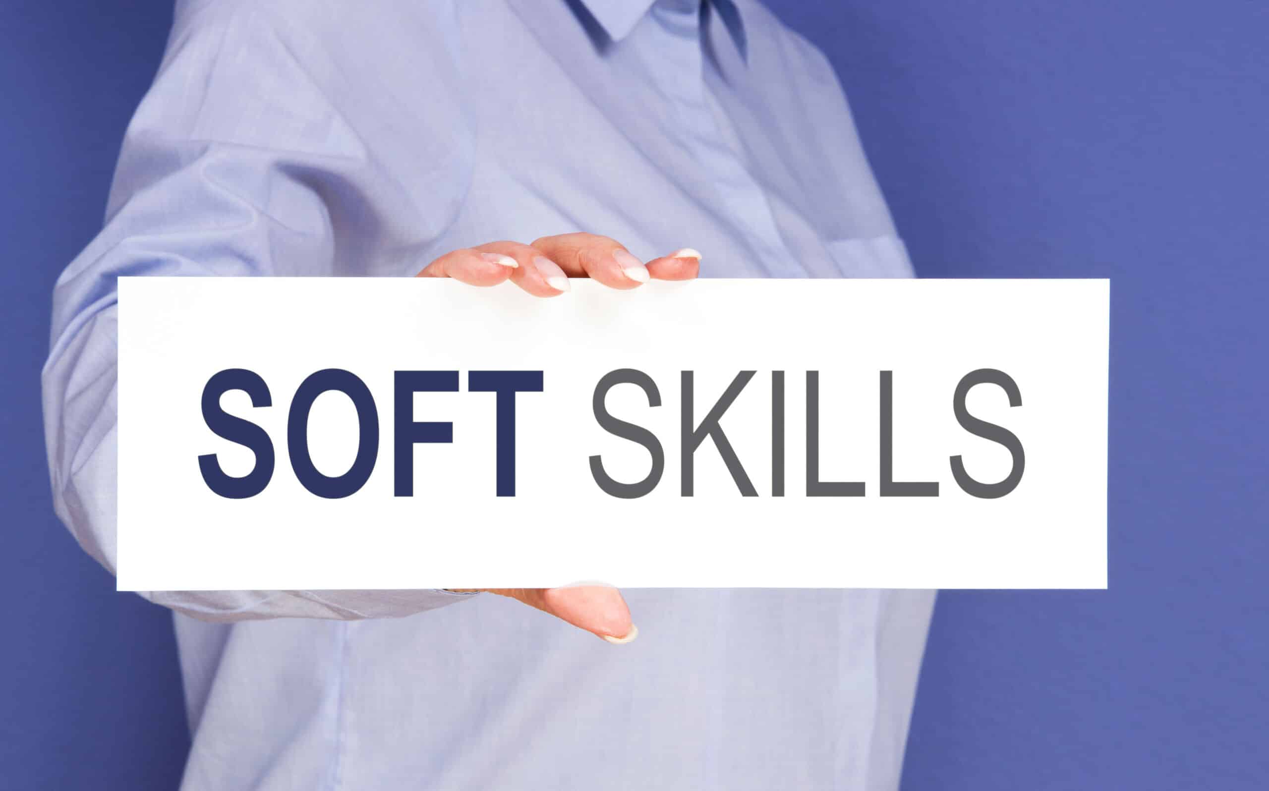 Person in einem hellblauen Hemd, die ein weißes Schild mit der Aufschrift "SOFT SKILLS" vor einem einfarbig blauen Hintergrund hält, um die Bedeutung der Rhetorik für Führungskräfte hervorzuheben.