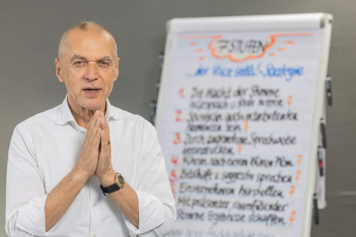 Ein älterer Mann im weißen Hemd steht vor einem verschwommenen Flipchart mit handschriftlichen Notizen und hält die Hände vor der Brust zusammen - bereit, seine Erkenntnisse über Rhetorik für Führungskräfte zu vermitteln.