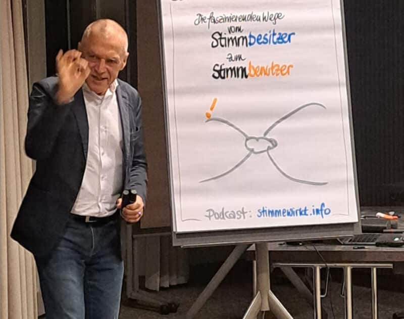 Ein Mann im Anzug gestikuliert neben einem Flipchart mit deutschem Text und einem Diagramm, das zeigt, wie man Überzeugen und Wirkung entfaltet, während er bei einer Indoor-Veranstaltung präsentiert.