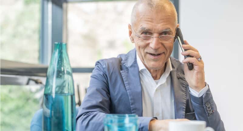 Ein älterer Mann in einem blauen Blazer und mit Brille telefoniert mit einem Smartphone an einem Tisch mit blauen Gläsern und einer klaren Wasserflasche.