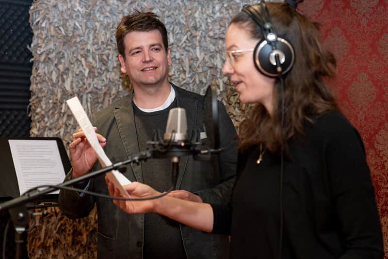Ein Mann mit einem Skript in der Hand steht neben einer Frau mit Kopfhörern, die in einem Tonstudio während ihrer Basisausbildung in Mikrofonsprechen in ein Mikrofon spricht.