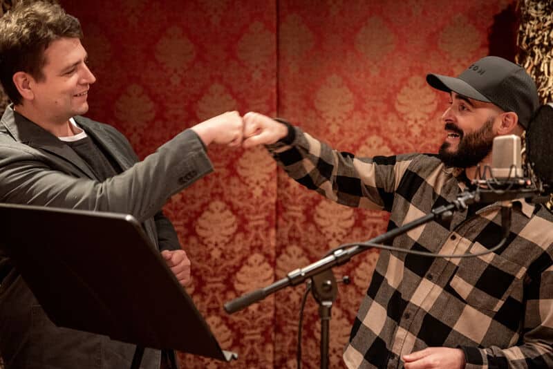 Zwei Männer stehen in einem Aufnahmestudio und geben sich einen Faustschlag, während sie vor einem gemusterten roten Hintergrund mit Mikrofon und Notenständer ihr Können beim Mikrofonsprechen unter Beweis stellen.