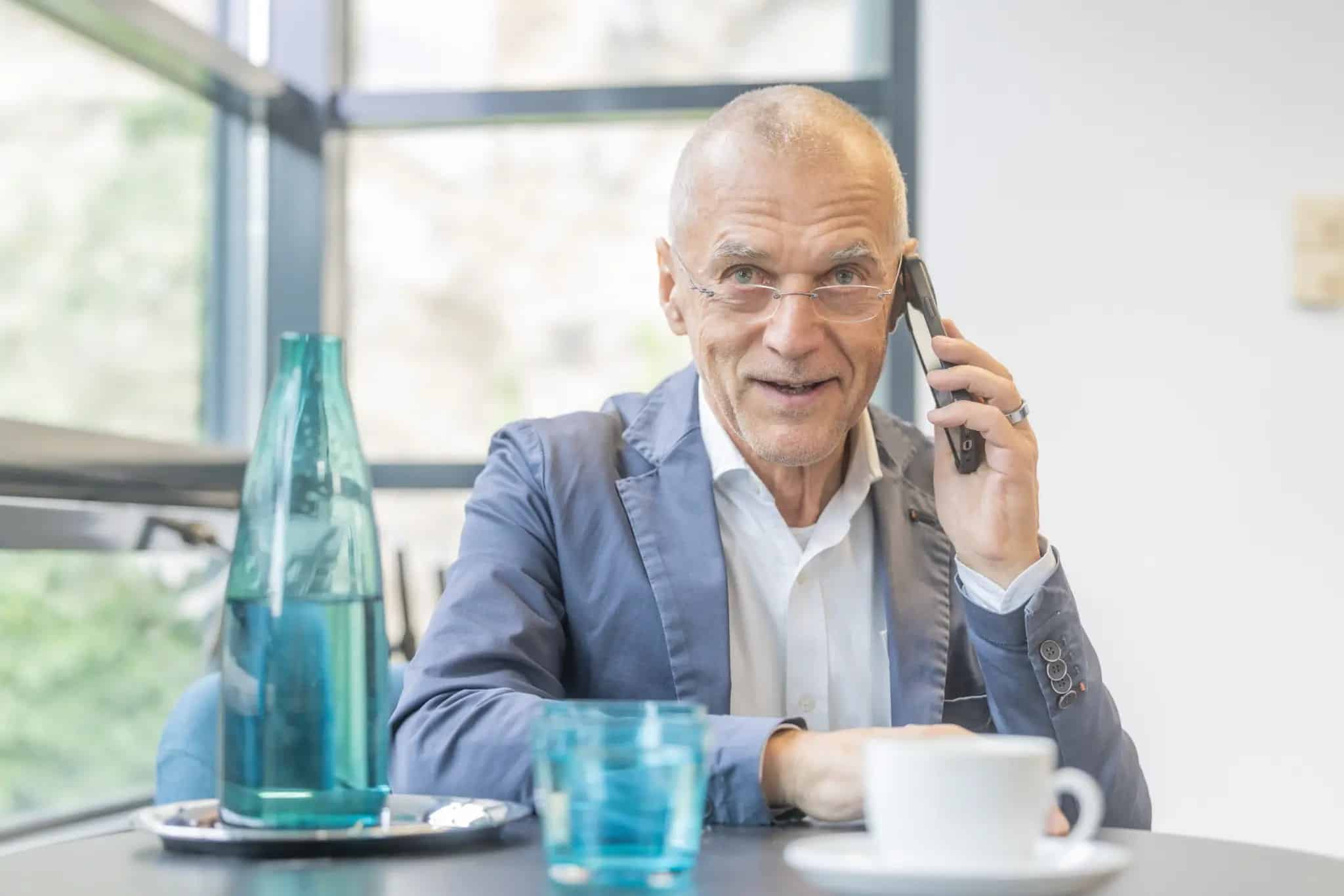 Ein älterer Mann im Anzug sitzt an einem Tisch mit einer Glasflasche, einem Glas und einer Tasse und spricht in einer hellen, modernen Umgebung mit einem Smartphone.