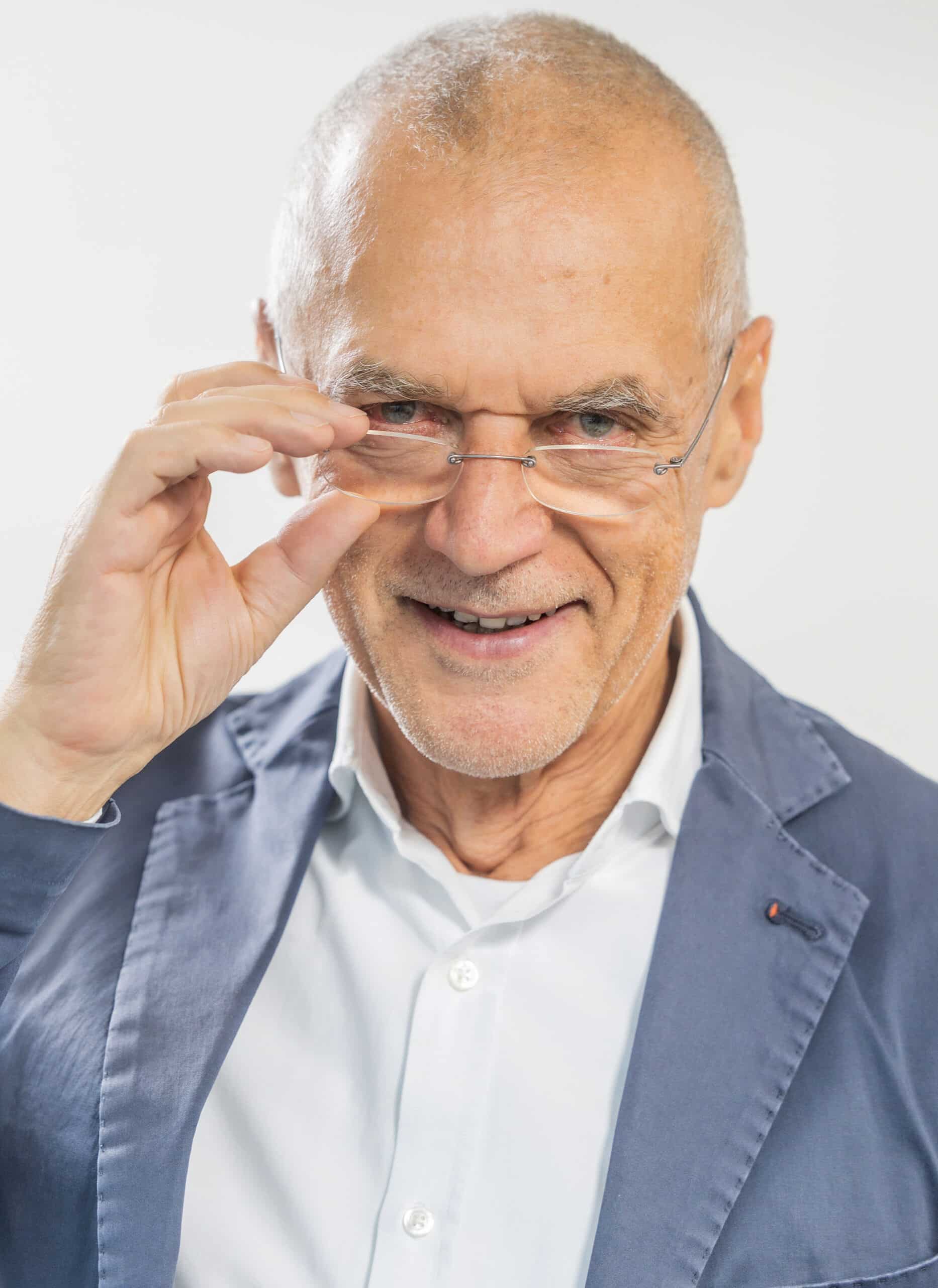 Arno Fischbacher, ein älterer Mann mit kurzen grauen Haaren, der eine Brille, ein weißes Hemd und einen blauen Blazer trägt; er lächelt, während er seine Brille zurechtrückt und in die Kamera blickt.