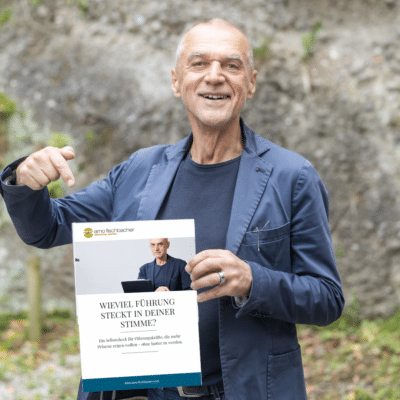 Ein Mann in einem blauen Blazer lächelt und zeigt auf eine Broschüre, die er in der Hand hält und auf der auf Deutsch „Wieviel Führung steckt in deiner Stimme?“ steht.