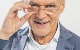 Ein älterer Mann in einem blauen Blazer und einem weißen Hemd rückt seine Brille zurecht und lächelt vor einem schlichten Hintergrund in die Kamera, als würde er sich auf eine offizielle Presseinformation vorbereiten.