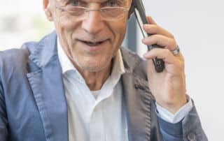 Ein älterer Mann mit Brille und Blazer telefoniert an einem Tisch, auf dem eine Kaffeetasse und ein blaues Glas stehen, mit seinem Smartphone und scheint gerade ein wichtiges Pressegespräch zu führen.