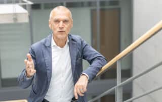 Ein Mann in einem blauen Blazer und einem weißen Hemd steht auf einer Treppe und gestikuliert mit einer Hand, als würde er eine Presseinformation vortragen – in einem modernen Gebäude mit Glaswänden und einem Metallgeländer.