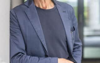 Ein älterer Mann in blauem Blazer und Jeans steht in einem Raum und lächelt in die Kamera, während er eine Hand in der Hosentasche hat, als wäre er bereit für eine offizielle Pressekonferenz.