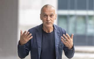 Ein kahlköpfiger Mann in einem marineblauen Blazer gestikuliert mit beiden Händen, während er draußen vor einem modernen Gebäude mit großen Fenstern steht, als wolle er bei einer Pressekonferenz wichtige Punkte hervorheben.
