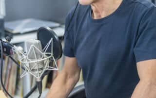 Ein Mann mit Kopfhörern sitzt an einem Schreibtisch mit Mikrofon, Tastatur und Maus und scheint in einem Studio eine Pressemitteilung aufzunehmen oder zu übertragen.