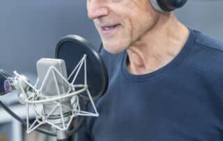 Ein älterer Mann mit Kopfhörern spricht in einem Tonstudio in ein Mikrofon und scheint an einer Radiosendung oder einem Podcast teilzunehmen, während er eine Pressemitteilung vorliest.