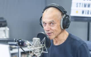 Ein kahlköpfiger Mann mit Kopfhörern spricht in ein Studiomikrofon und verliest eine Pressemitteilung; im Hintergrund ist ein unscharfes Bild zu sehen, und ein Buchstabenbrett ist teilweise sichtbar.