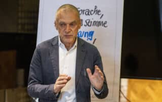 Ein Mann im Anzug gestikuliert, während er vor einem Flipchart mit handschriftlichen Notizen spricht und im Rahmen einer Presseinformation wichtige Details vorstellt.