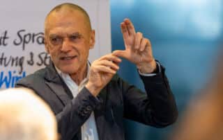 Ein Mann im Anzug gestikuliert beim Sprechen mit den Händen und steht dabei vor einer Weißwandtafel, auf der teilweise Schriftzüge zu sehen sind, als würde er seinem Publikum eine wichtige Pressemitteilung vortragen.